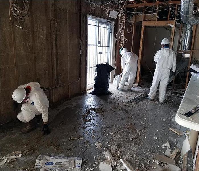 office with water damaged floor covered in debris and 3 employees in white coveralls cleaning